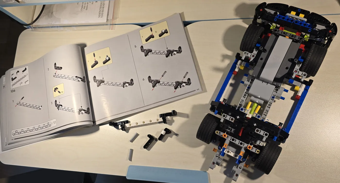 Schreibtisch mit aufgeschlagener Bauanleitung, einzelnen Bauteilen und dem halb fertigen Chassis des Fahrzeugs.